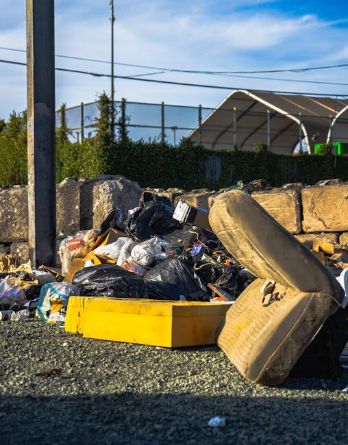 An outdoor scene showing a pile of mixed waste and refuse bins on a gravel surface, with several black plastic bin bags, a yellow plastic crate, a large discarded shoe with a beige fabric cover, and various smaller bits of rubbish scattered around. Behind the waste, there is a low stone wall composed of rough, weathered stones, and in the background, a metal pole with power lines, green foliage, and a partially obscured modern building with a curved, metallic roof. The environment appears to be an informal rubbish collection point or an area where waste has been temporarily left. The lighting suggests daytime, with clear skies overhead, highlighting the textures of the waste materials and surroundings. Waste Clearance Roehampton manages waste collection in the area, providing private options for rubbish removal outside of council services, with this scene illustrating the need for proper disposal or on-site clearance services for similar deposits of refuse.