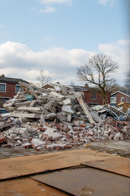 A large pile of demolished building materials, including broken concrete slabs, bricks, and metal debris, is situated on a construction site with exposed ground and wooden boarding in the foreground. The rubble is irregularly stacked, with some sections leaning and others scattered across the area. In the background, there are residential houses with dark brick facades and pitched roofs, set against a partly cloudy sky with some trees without leaves. The scene appears to be a site prepared for waste clearance or site clearing activities, consistent with the services offered by Waste Clearance Roehampton for independent or private waste removal related to building demolition or refurbishment projects.