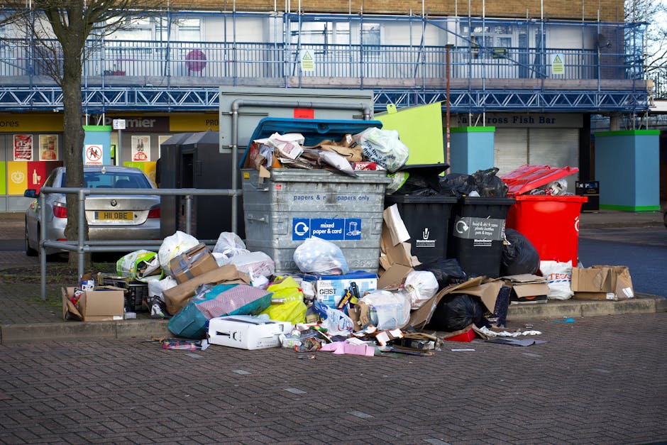 An outdoor scene showing a pile of mixed waste and refuse bins on a gravel surface, with several black plastic bin bags, a yellow plastic crate, a large discarded shoe with a beige fabric cover, and various smaller bits of rubbish scattered around. Behind the waste, there is a low stone wall composed of rough, weathered stones, and in the background, a metal pole with power lines, green foliage, and a partially obscured modern building with a curved, metallic roof. The environment appears to be an informal rubbish collection point or an area where waste has been temporarily left. The lighting suggests daytime, with clear skies overhead, highlighting the textures of the waste materials and surroundings. Waste Clearance Roehampton manages waste collection in the area, providing private options for rubbish removal outside of council services, with this scene illustrating the need for proper disposal or on-site clearance services for similar deposits of refuse.