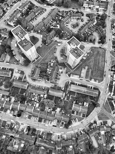 An aerial black-and-white photograph shows a densely populated urban area featuring a mix of residential houses and larger multi-storey buildings. In the foreground, rows of terraced houses with pitched roofs and small front gardens are visible, with parked cars lining the narrow streets. The middle ground displays two prominent high-rise buildings with flat roofs, surrounded by open green spaces and pathways, indicating a possible communal or recreational area. Behind these high-rise blocks, there is a cluster of smaller, tightly packed residential buildings with varied rooflines and cluttered backyards. The scene is lit by natural daylight, with visible shadows cast by the buildings and trees. The overall layout reflects a typical urban environment with closely situated dwellings and minimal open space, aligned with areas where private waste collection or on-site clearance might be conducted by services such as Waste Clearance Roehampton for efficient rubbish removal and waste management.