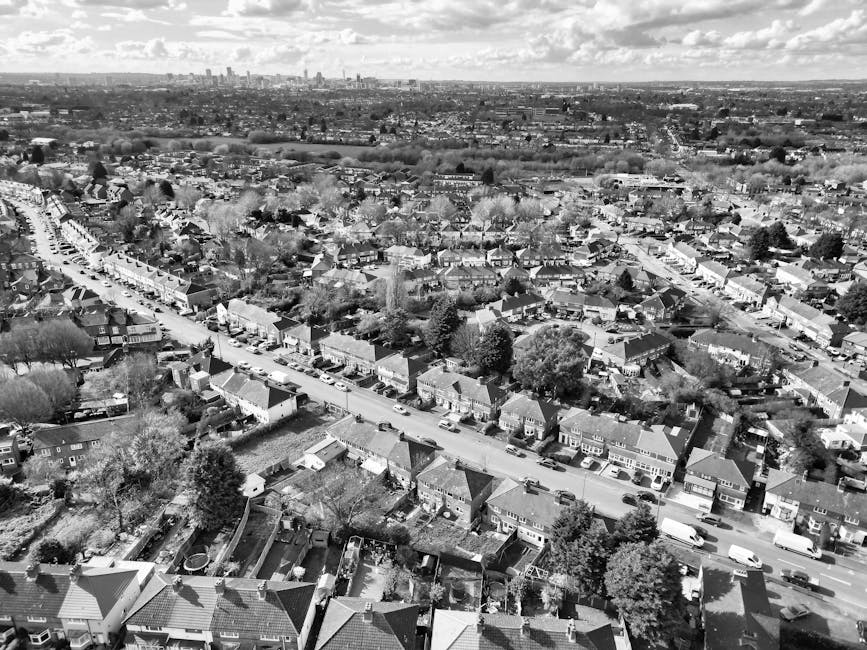 An aerial black-and-white photograph shows a densely populated urban area featuring a mix of residential houses and larger multi-storey buildings. In the foreground, rows of terraced houses with pitched roofs and small front gardens are visible, with parked cars lining the narrow streets. The middle ground displays two prominent high-rise buildings with flat roofs, surrounded by open green spaces and pathways, indicating a possible communal or recreational area. Behind these high-rise blocks, there is a cluster of smaller, tightly packed residential buildings with varied rooflines and cluttered backyards. The scene is lit by natural daylight, with visible shadows cast by the buildings and trees. The overall layout reflects a typical urban environment with closely situated dwellings and minimal open space, aligned with areas where private waste collection or on-site clearance might be conducted by services such as Waste Clearance Roehampton for efficient rubbish removal and waste management.