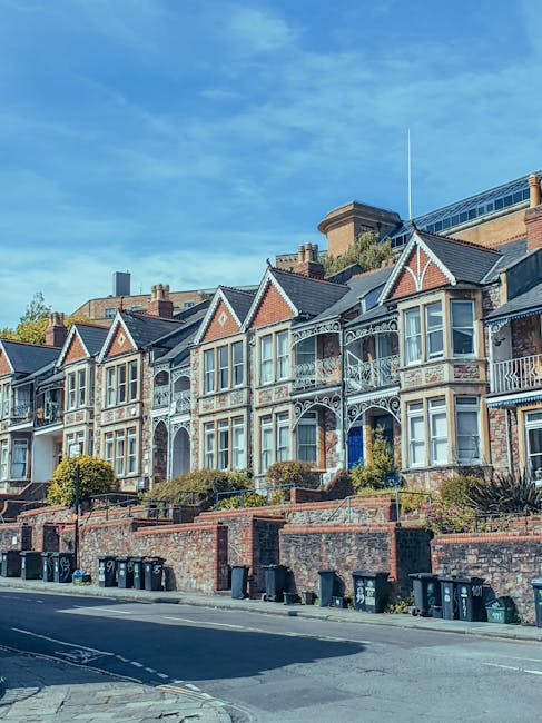 A row of Victorian-style terraced houses with decorative ironwork and bay windows, set on a raised brick terrace with small landscaped front gardens. The houses are clad in a combination of brick and painted finishes, with slate roofs and gabled facades. In the foreground, a street with several black wheelie bins is visible along the pavement. The houses are situated in an urban residential area, with a modern building partially visible in the background behind the rooftops, and a clear blue sky overhead. The scene is well-lit with natural daylight, emphasizing the architectural details and the tidy appearance of the street, relating to private property waste management and alternative rubbish removal options like local collection services managed by companies such as Waste Clearance Roehampton.
