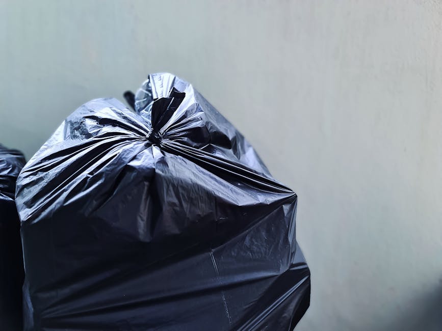 Close-up view of black plastic rubbish bags filled with waste, tightly packed and tied at the top, resting against a plain, light-colored wall. The bags are made of shiny, crinkled plastic material, with visible creases and folds. The scene suggests an indoor or outdoor setting suitable for private waste collection or on-site clearance, as part of a rubbish removal service. The bags occupy the foreground, with minimal background detail, emphasizing their size and condition. This image exemplifies typical materials handled during waste clearance activities, aligning with private waste disposal options for household or commercial rubbish. The lighting is neutral, highlighting the reflective surface of the plastic and the texture of the waste, suitable for visual documentation of rubbish collection tasks performed by Waste Clearance Roehampton or similar services in the area.